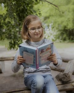 Ein Mädchen im Grundschulalter mit hellen Haaren und Brille, den Blick freundlich und konzentriert auf das Buch gerichtet, das sie in den Händen hält.