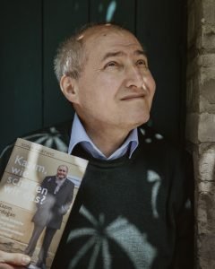Ein Mann mit grauen Haaren, den Blick eher ernst leicht nach oben gerichtet, mit einem Buch in der Hand, auf dessen Cover er selbst abgebildet ist.